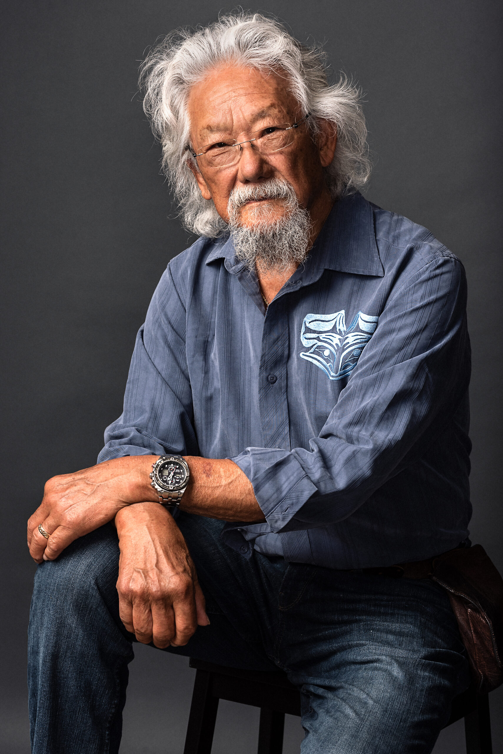 An elderly man with grey hair and a grey beard sits casually in a blue button-up shirt. 