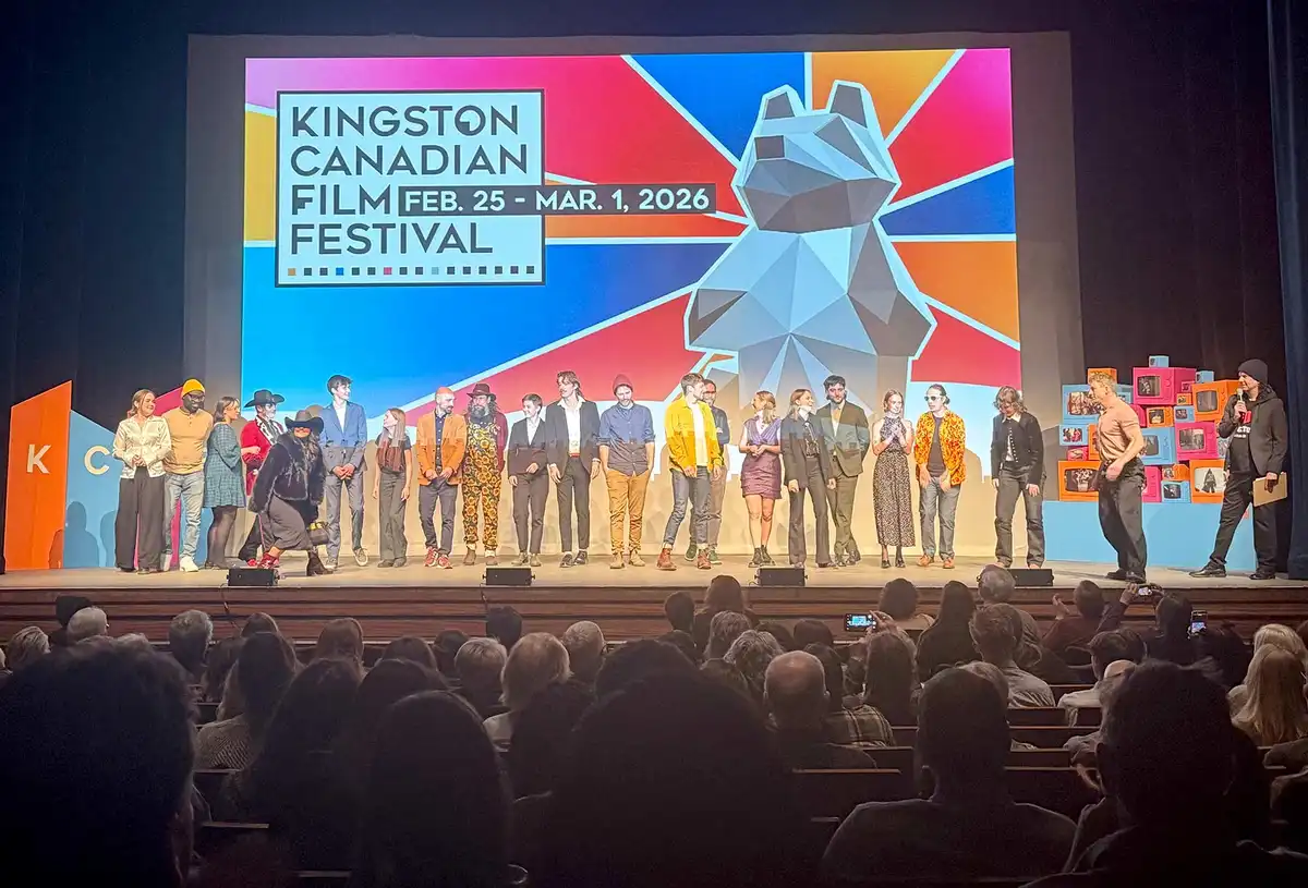 A group of people stand in a line on a stage. An audience of people looks up at that. A screen at the back of the stage says, "Kingston Canadian Film Festival" and "Feb. 25 - Mar. 1, 2026". The colours behind the text are bright lines with a silver figure.
