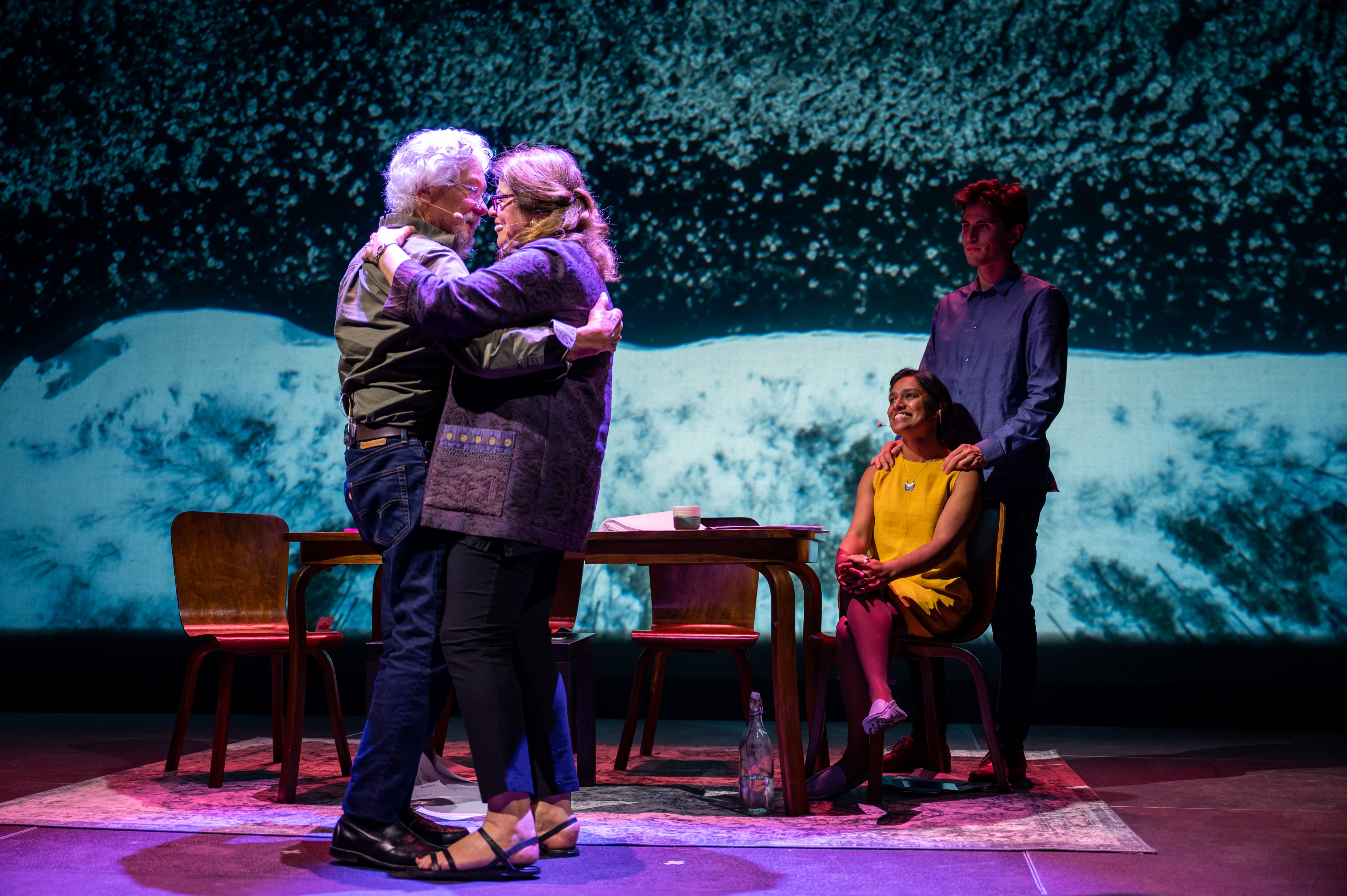 An older man and woman dance close together on stage. Further back, a younger man and woman watches them. 