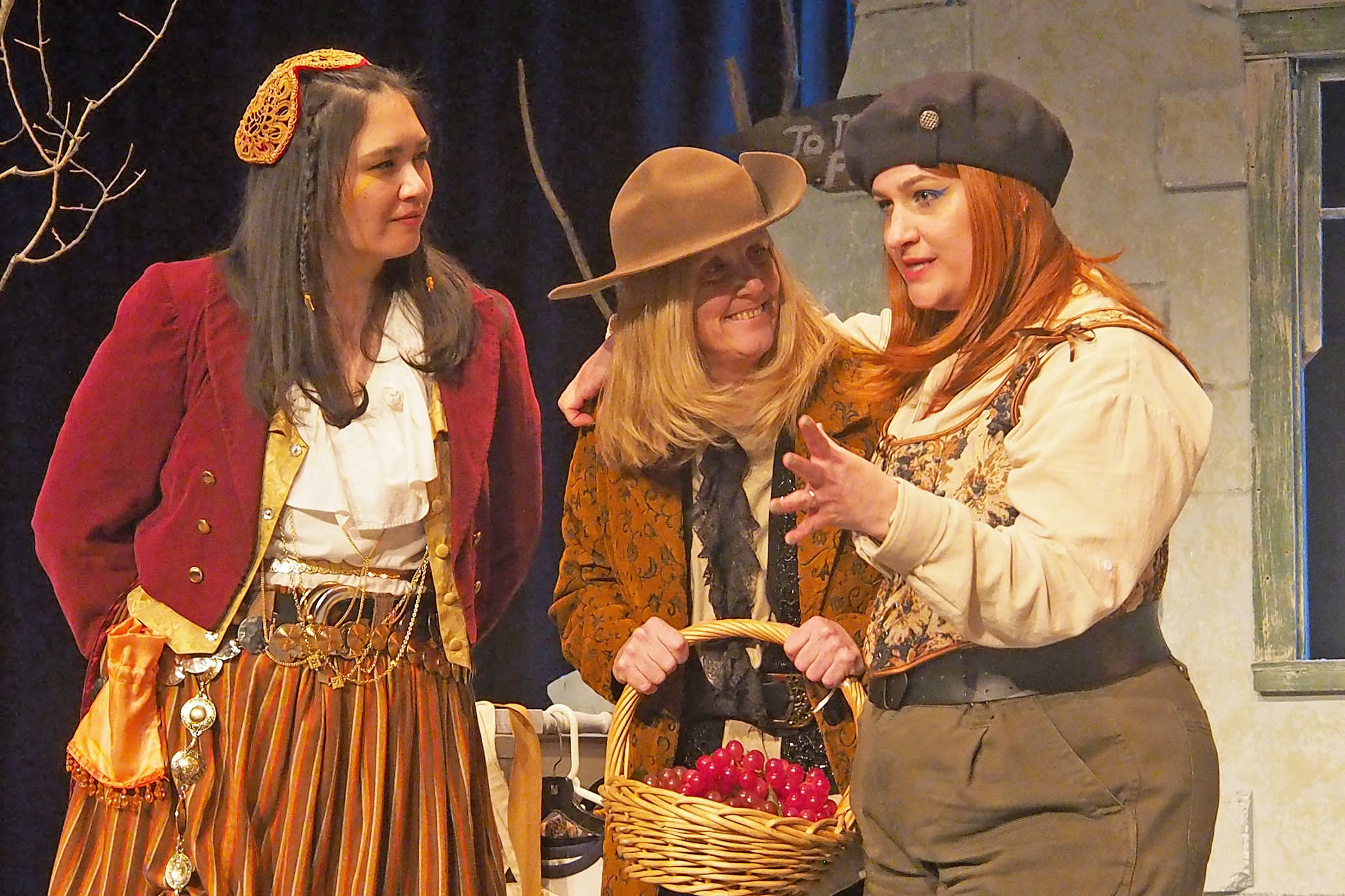Three actors on stage, the middle one holds a wicker basket while the one on the right speaks. 