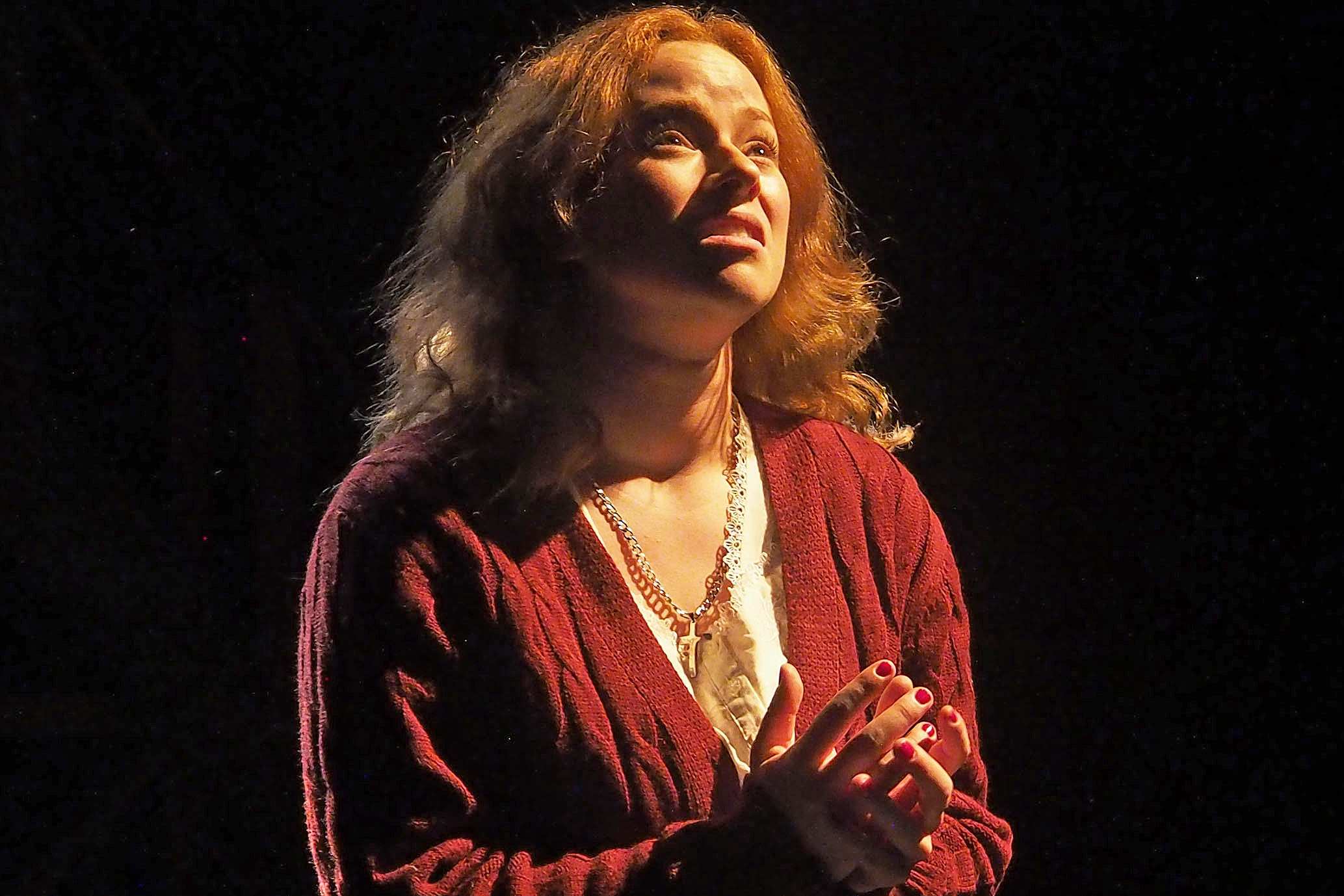 A medium shot of a woman with long hair wearing a red cardigan, hands clasped in prayer.
