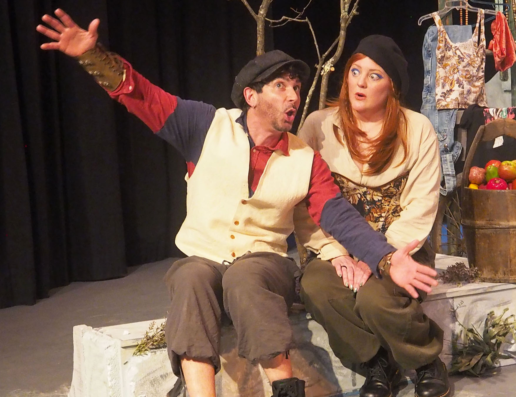 A man and woman sit on stage, the man spreads his arms wide apart. 