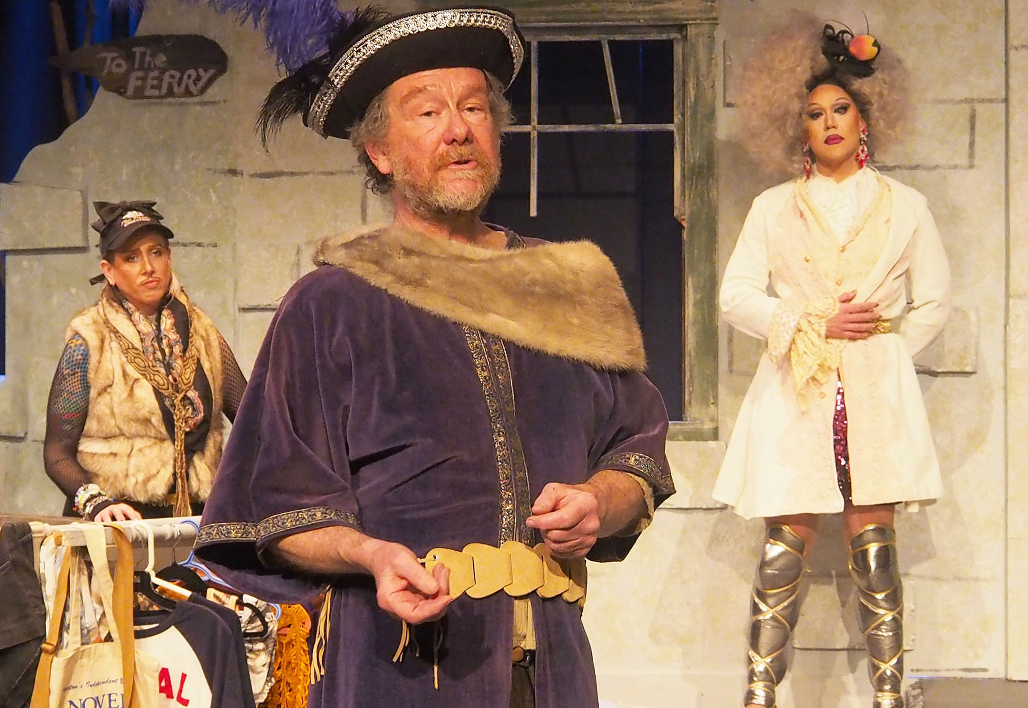 Two actors in the background. In the foreground, a man speaks wearing a bard's hat. 
