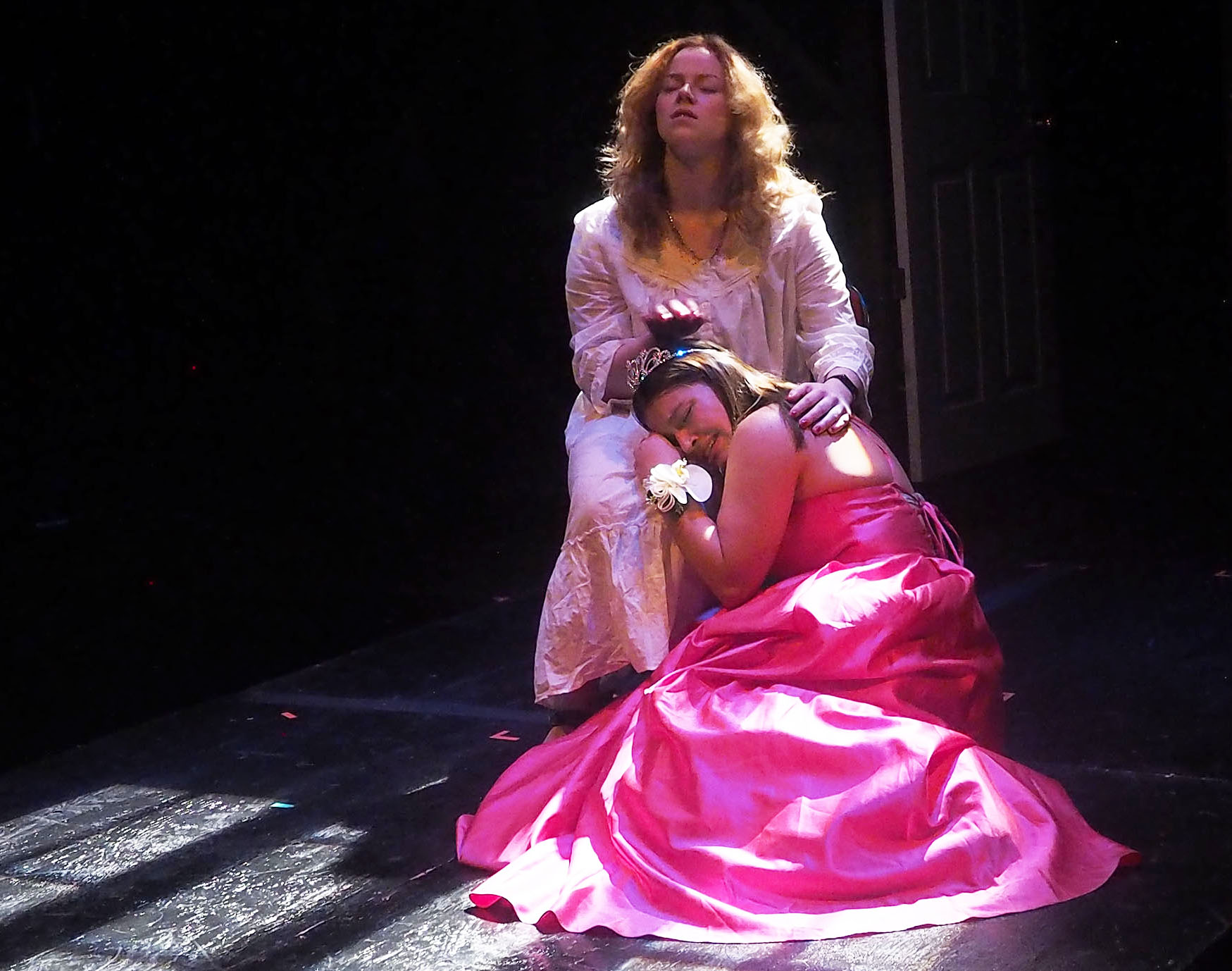 A woman with dark hair wearing a hot pink gown sits on the floor with her head in the lap of another woman with lighter hair. 