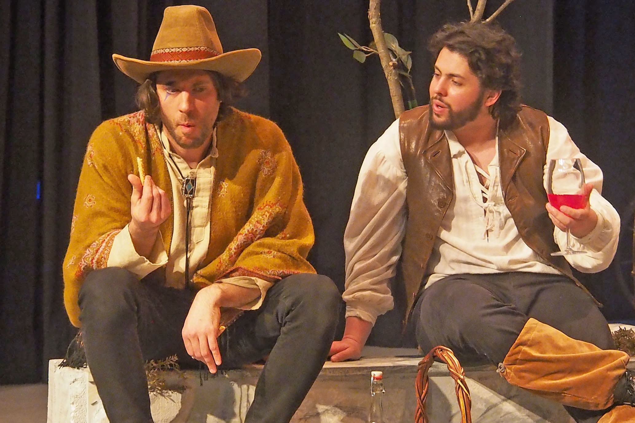 Two men sit on stage. On the left, he wears a cowboy hat, on the right, he wears a vest and holds a glass of something red. 