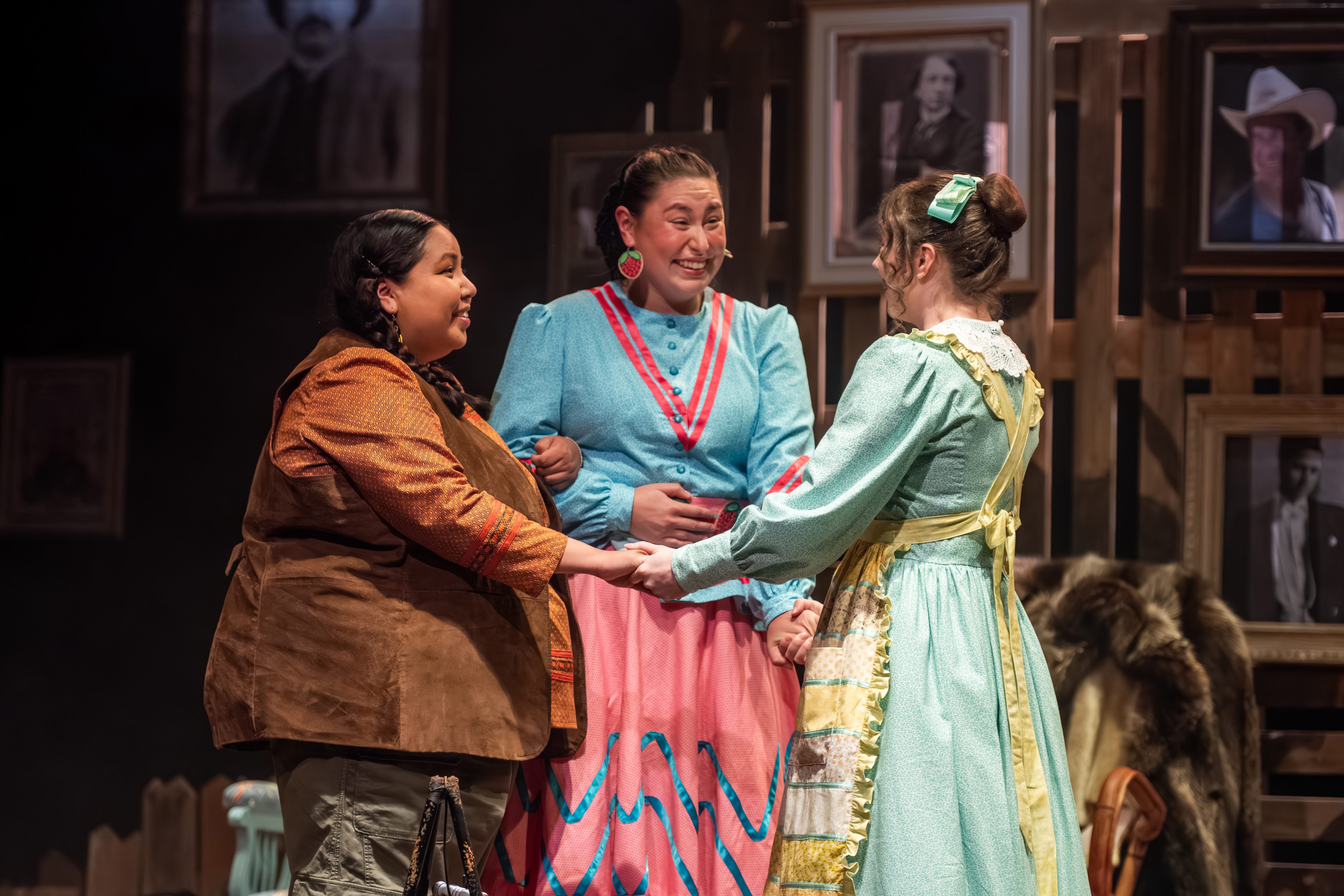 Three women stand together, holding each other and looking happy. Two wear Indigenous garments and the other wears a dress and apron. 