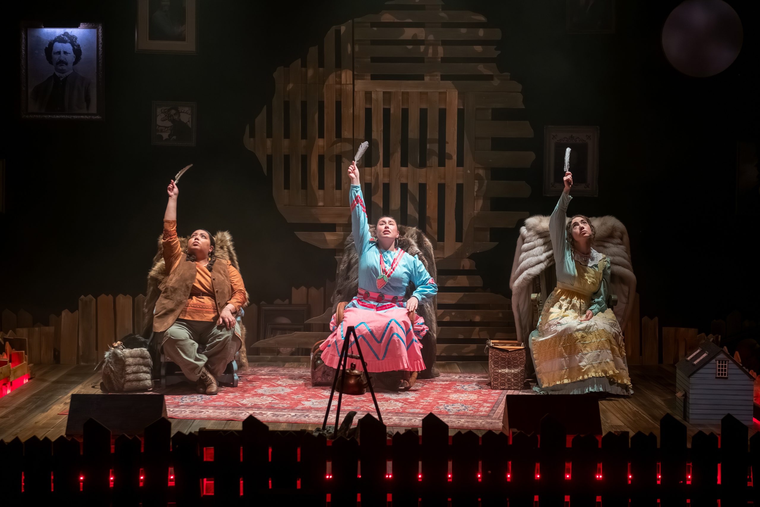 Three women performing. They each sit in a chair and raise an object to the sky. They are against a wooden backdrop, with their chairs atop a carpet. Two women wear Indigenous garments while a third wears a dress and apron.