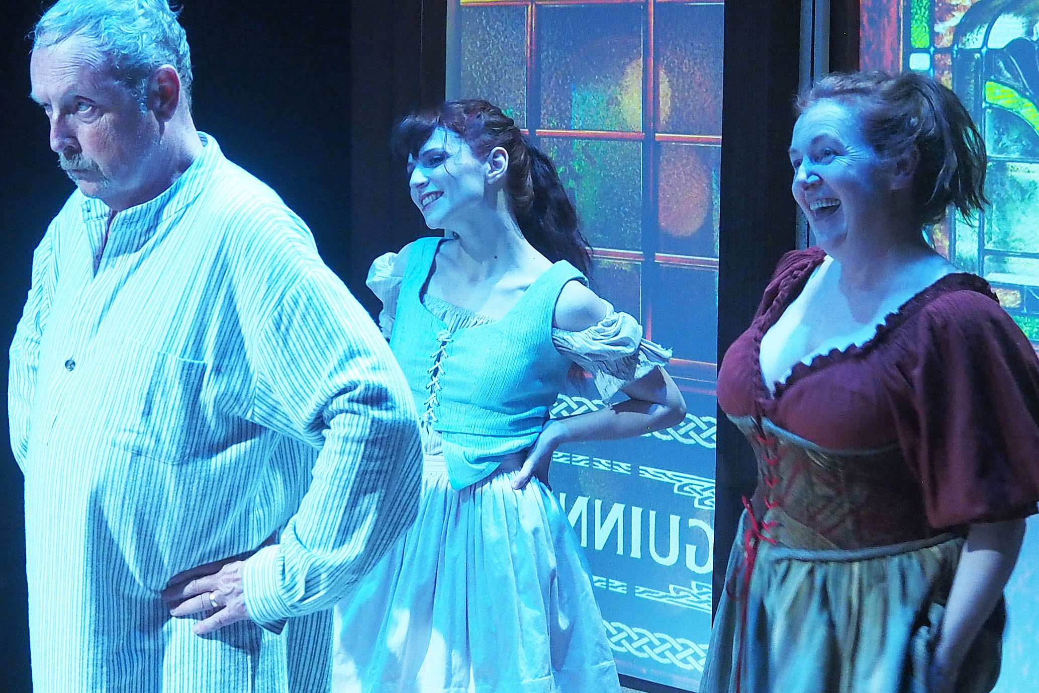 Three actors performing on stage. A man is in the middle, looking unimpressed with his hands on his hips. Two women in old-timey dresses stand behind him looking very happy. 