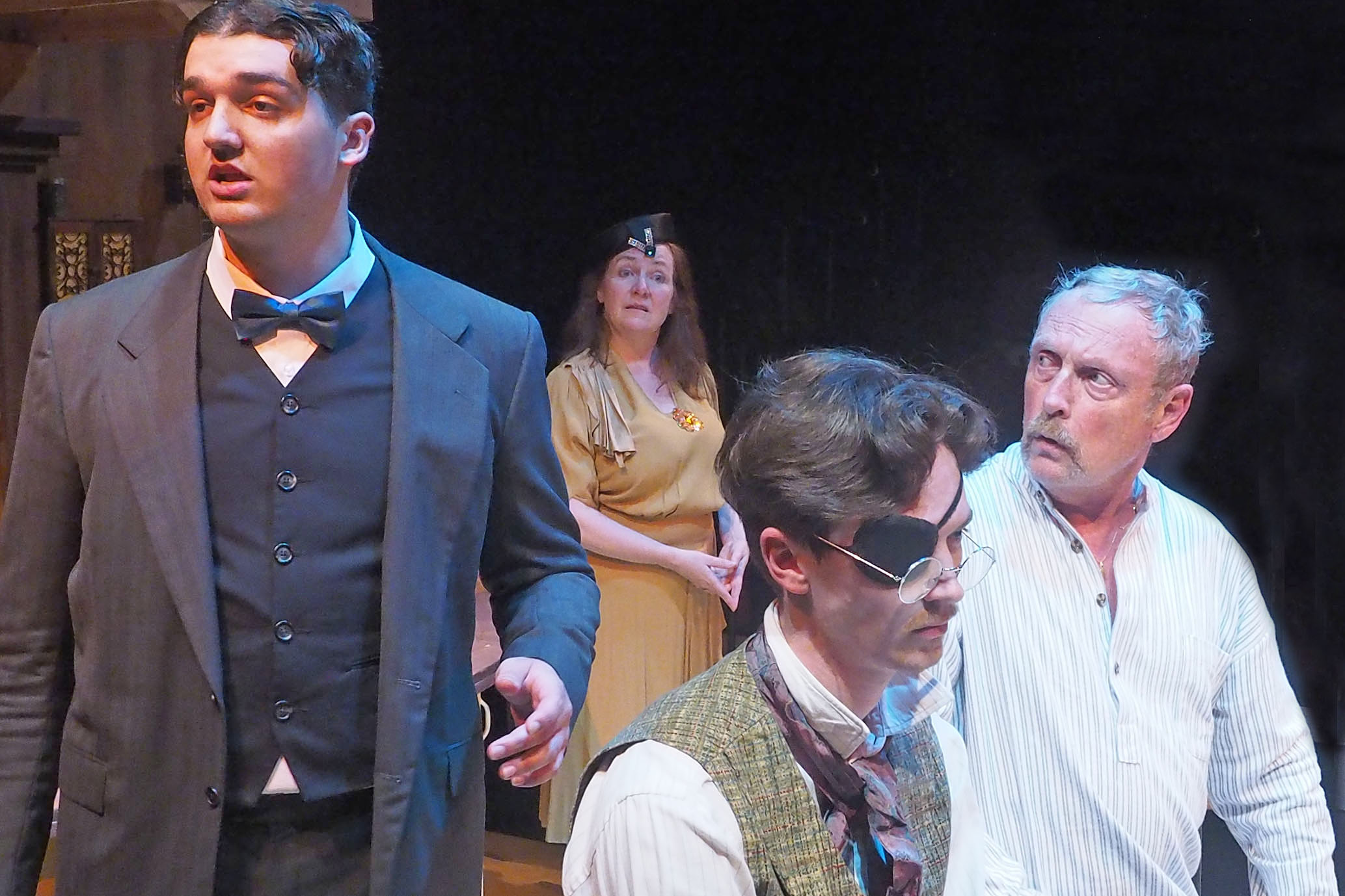 Four actors performing on stage. One wears a three-piece suit, another wears a plain shirt staring concernedly at the man in the suit, another wears an eyepatch looking in front of him, and a woman stands in the back, wearing a head piece of the 1920s. 