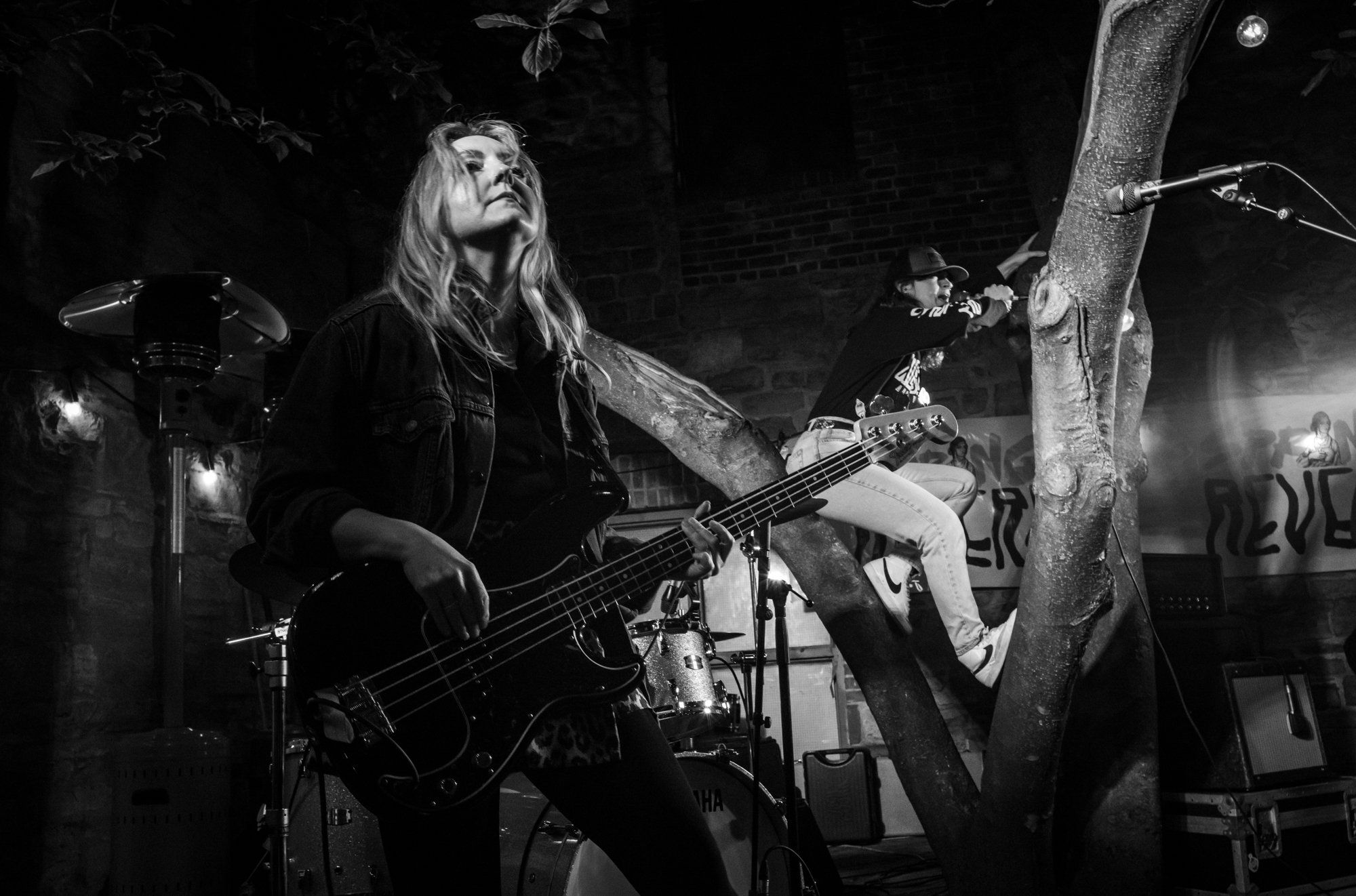A black and white image of a performer playing a guitar while atop an outdoor stage while another performer sings from the branches of a tree.