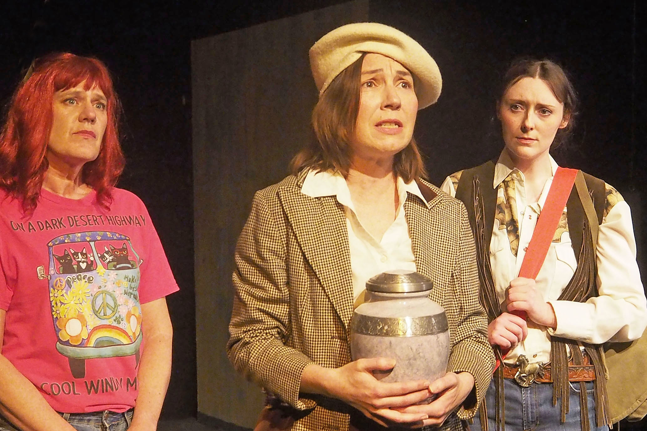 Three women look stoic. The one standing in the centre holds an urn. 