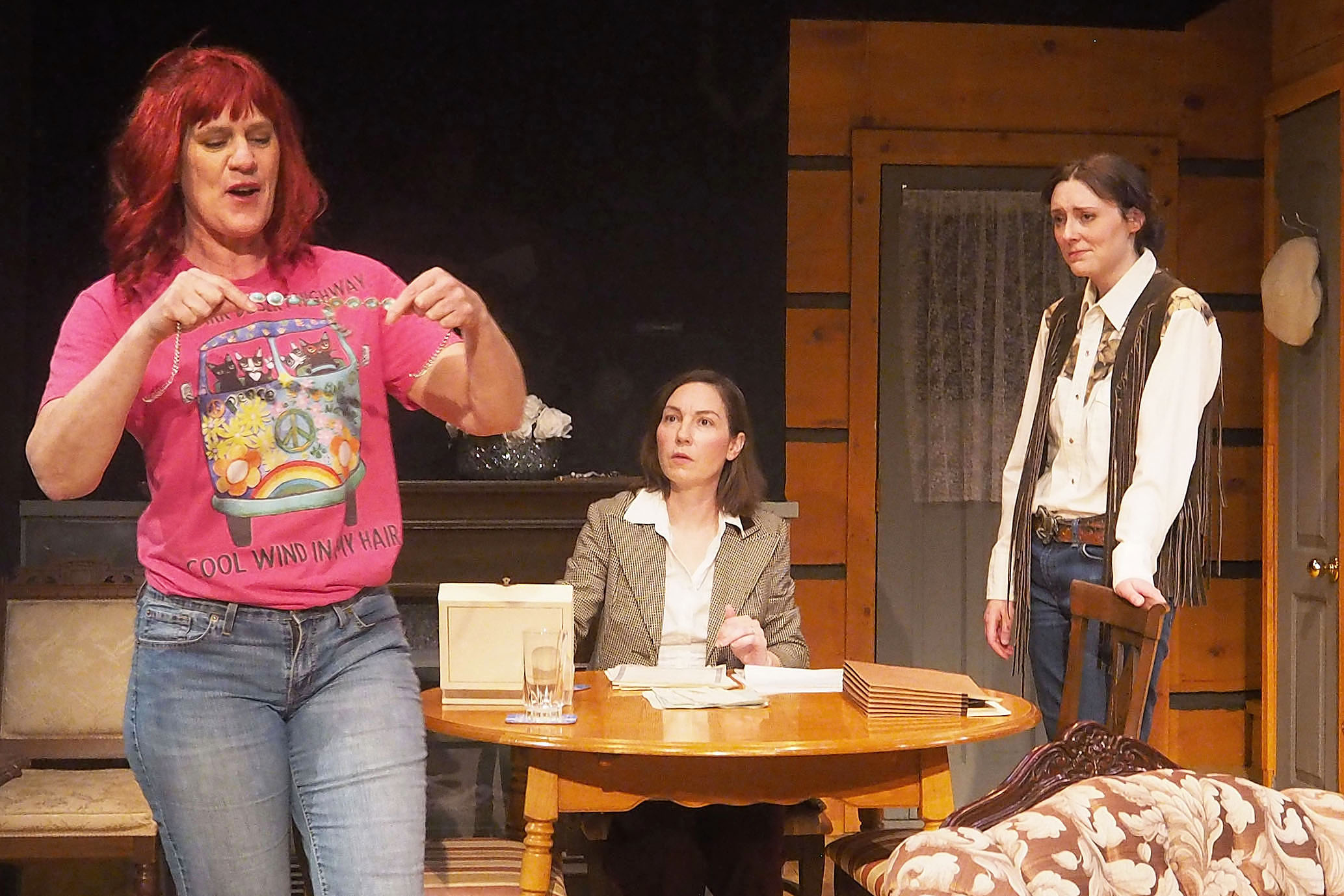 Three woman are around a kitchen table. One sits and two stand. One woman holds out a friendship bracelet. 
