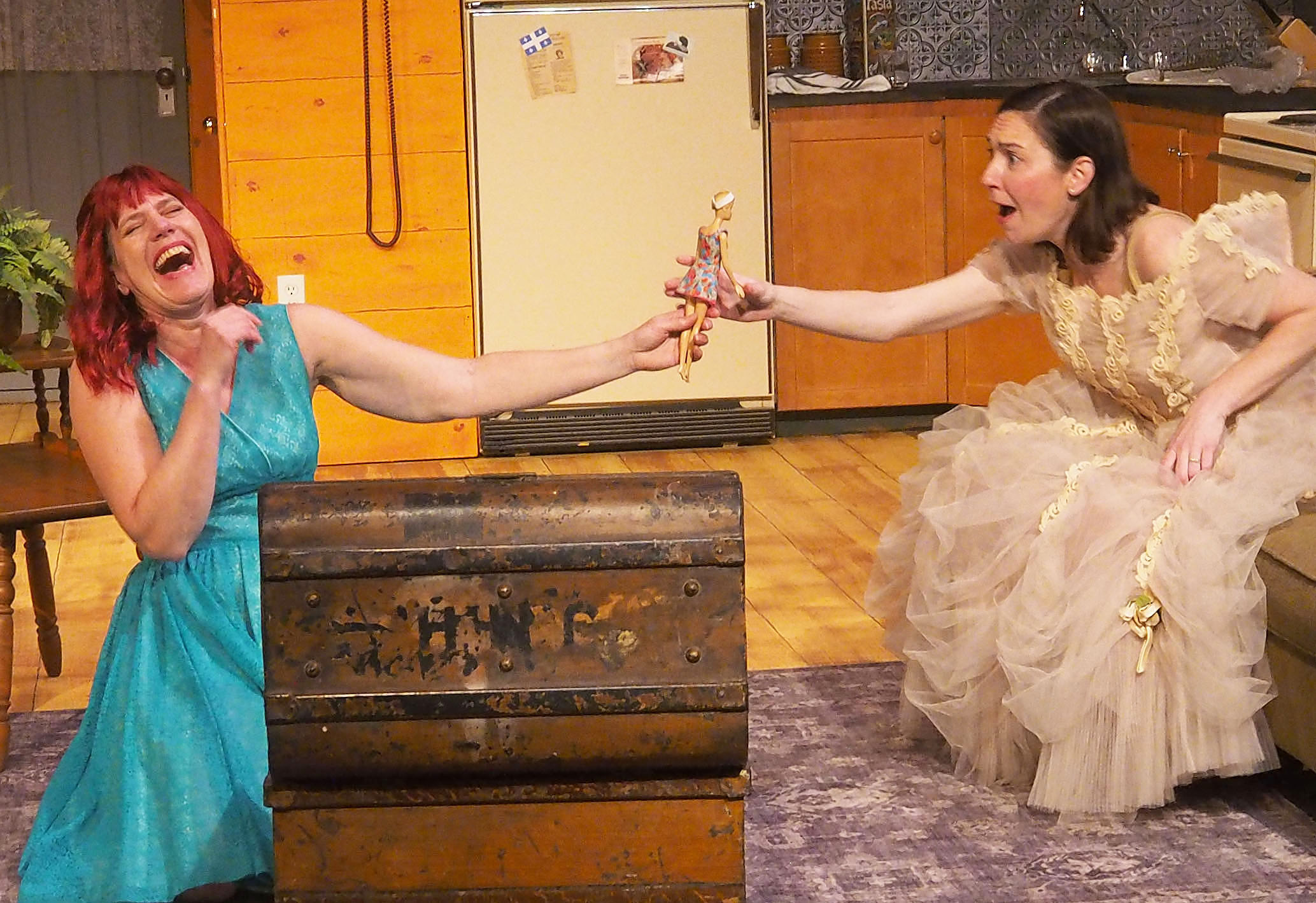 Two women hold a doll overtop a chest, laughing in the middle of a kitchen. 
