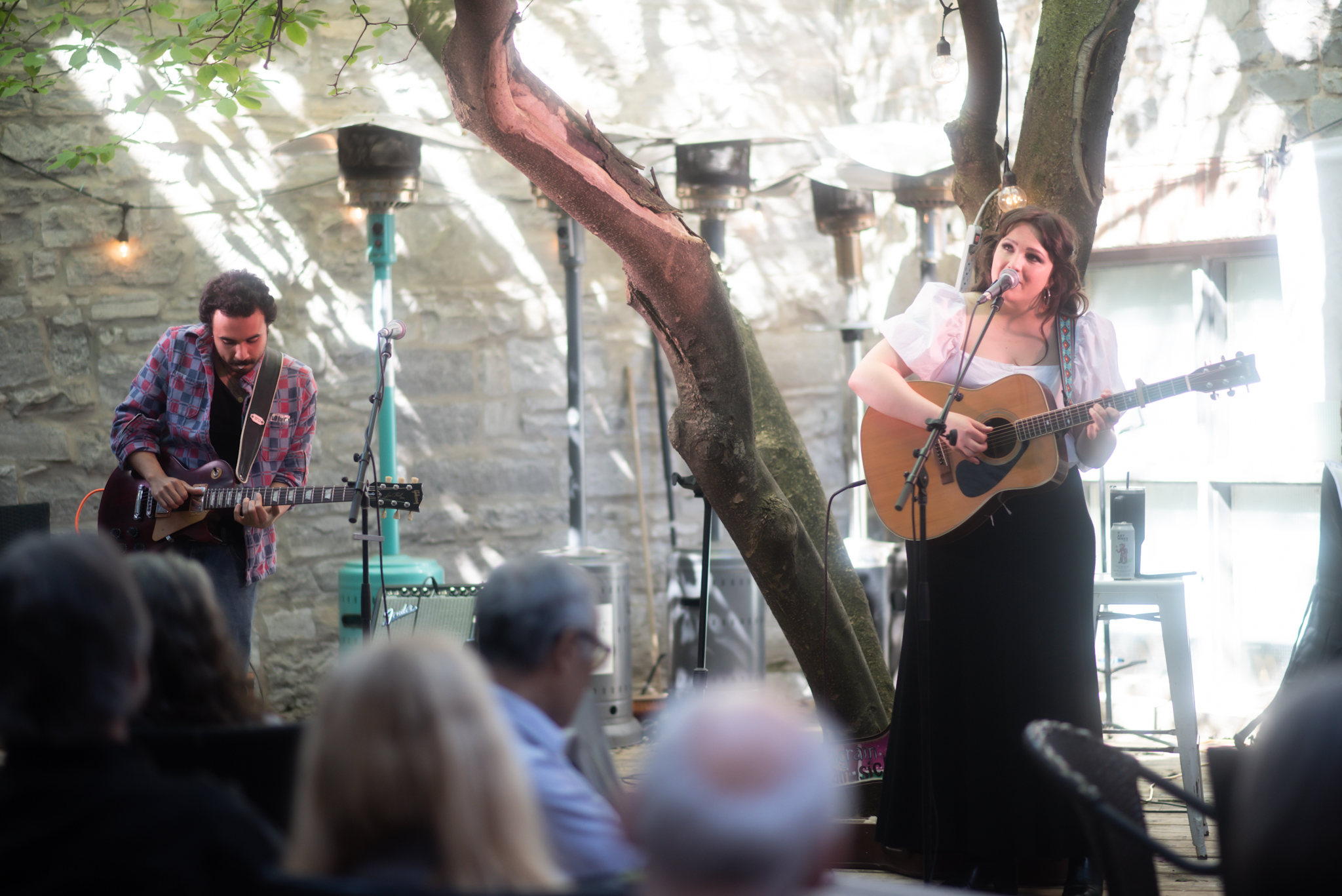 Two people performing on stage. A man appears in the back playing an electric guitar. A woman sings into a microphone and holds a guitar. 