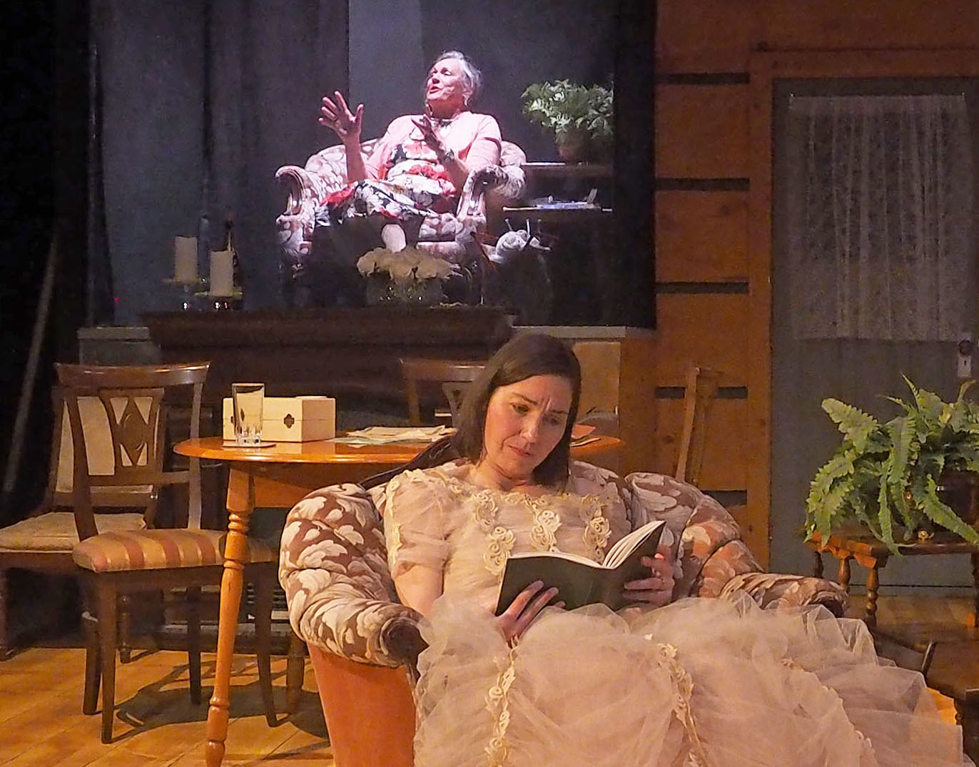 One woman reads in a sitting chair while another sits upstage with a light shining on her. They seem to be in two different scenes. 