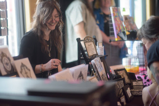 A vendor stands behind her booth smiling. On her table are framed drawings. 