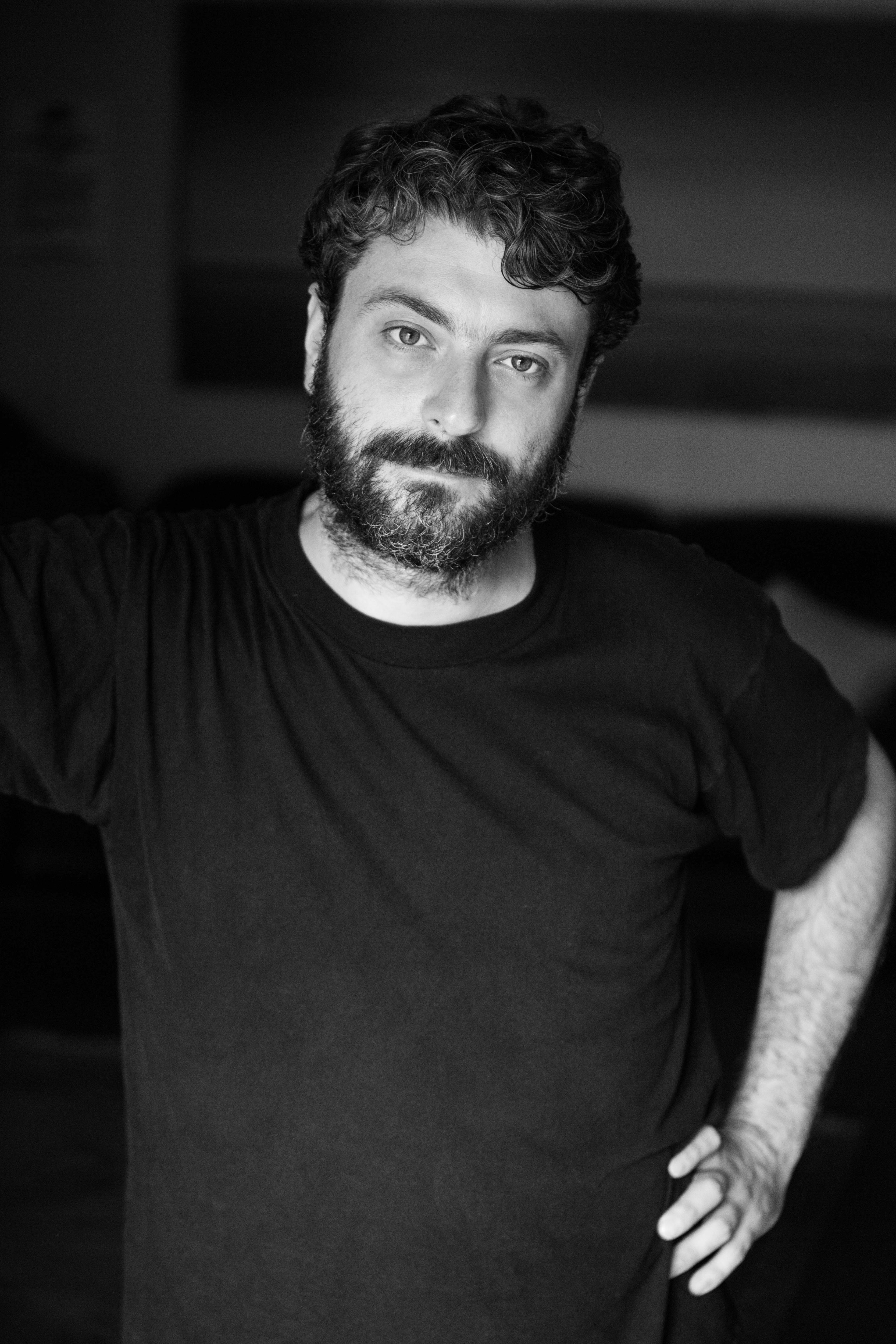 A white man with dark hair and beard looks at the camera in black and white.