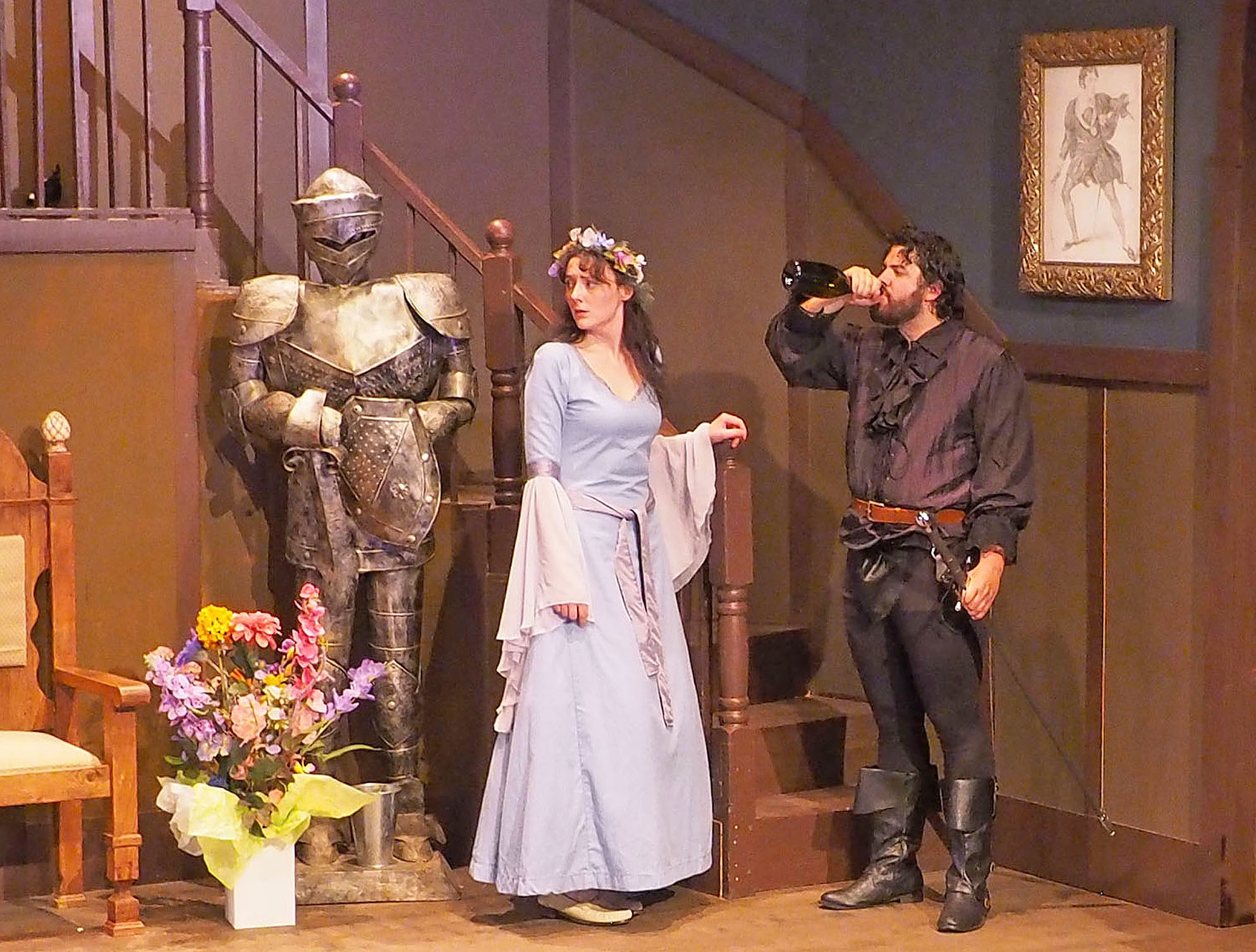 A woman in renaissance costume stands in the middle of the photograph, with a suit of armour to her right and a man drinking from a bottle of alcohol to her left.