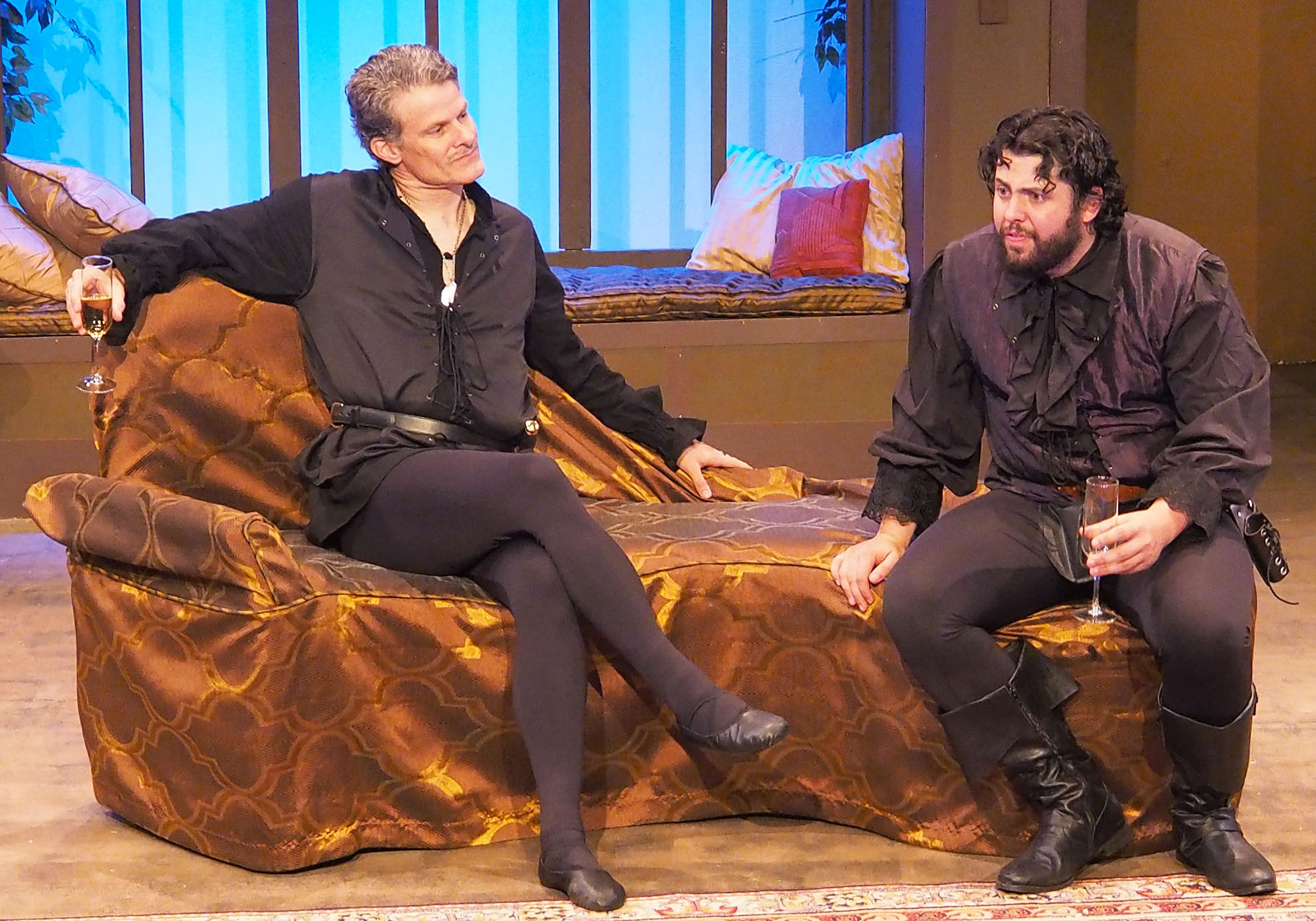 A photograph of two men sitting on a couch; both are relaxed, wearing renaissance costumes.