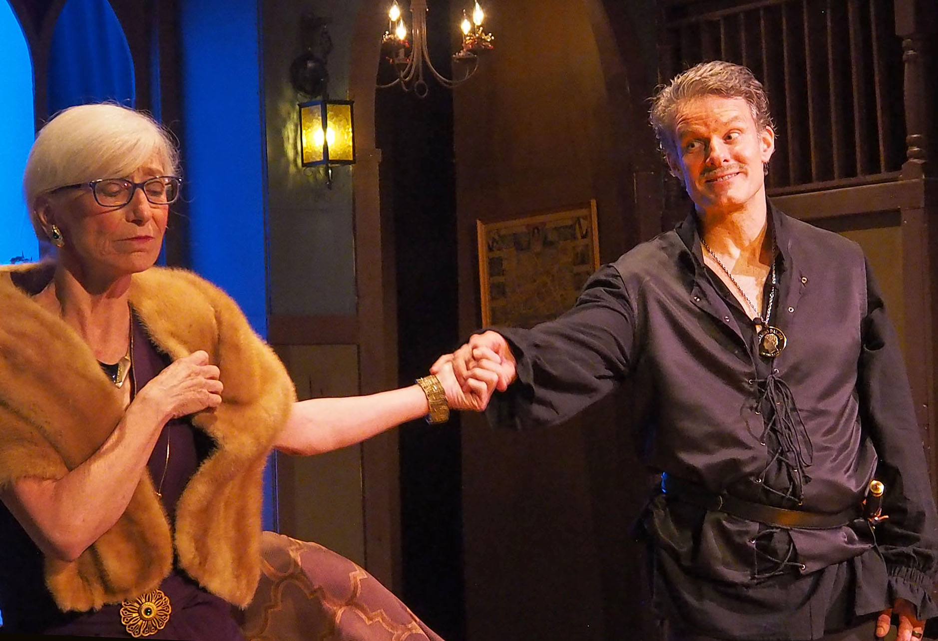 A photograph of a woman (left) and man (right). The woman appears distraught, wrapped in a fur coat with her eyes closed. The man reaches out and holds her hand with an energetic expression.