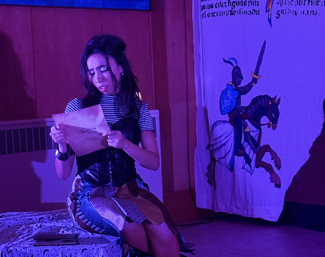 A performer in punk-style clothing sits on the edge of a bed onstage, reading a letter.