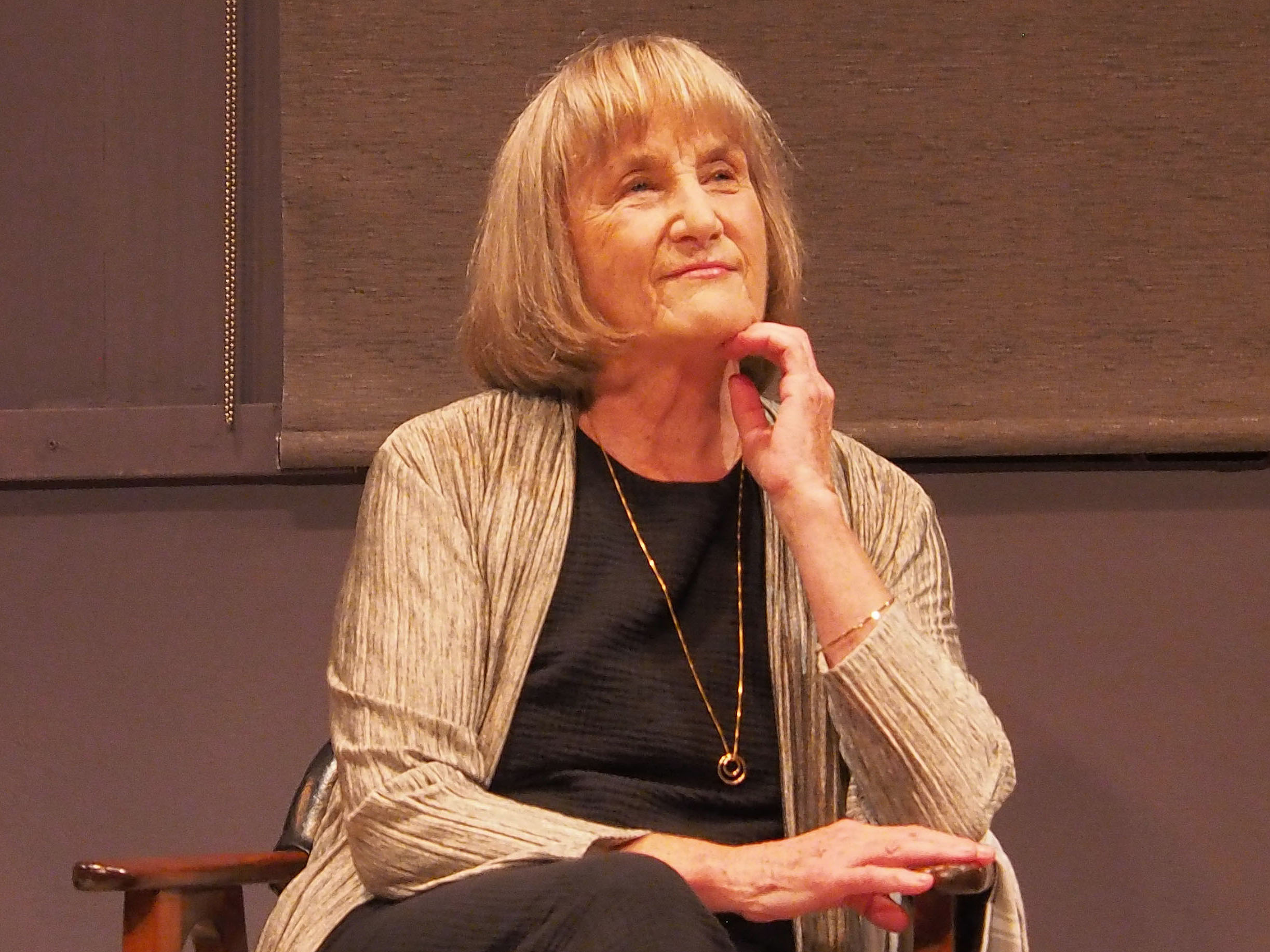 McBride, an older woman with a blonde bob haircut, seated, stares pensively into the distance with a hand beneath her chin.