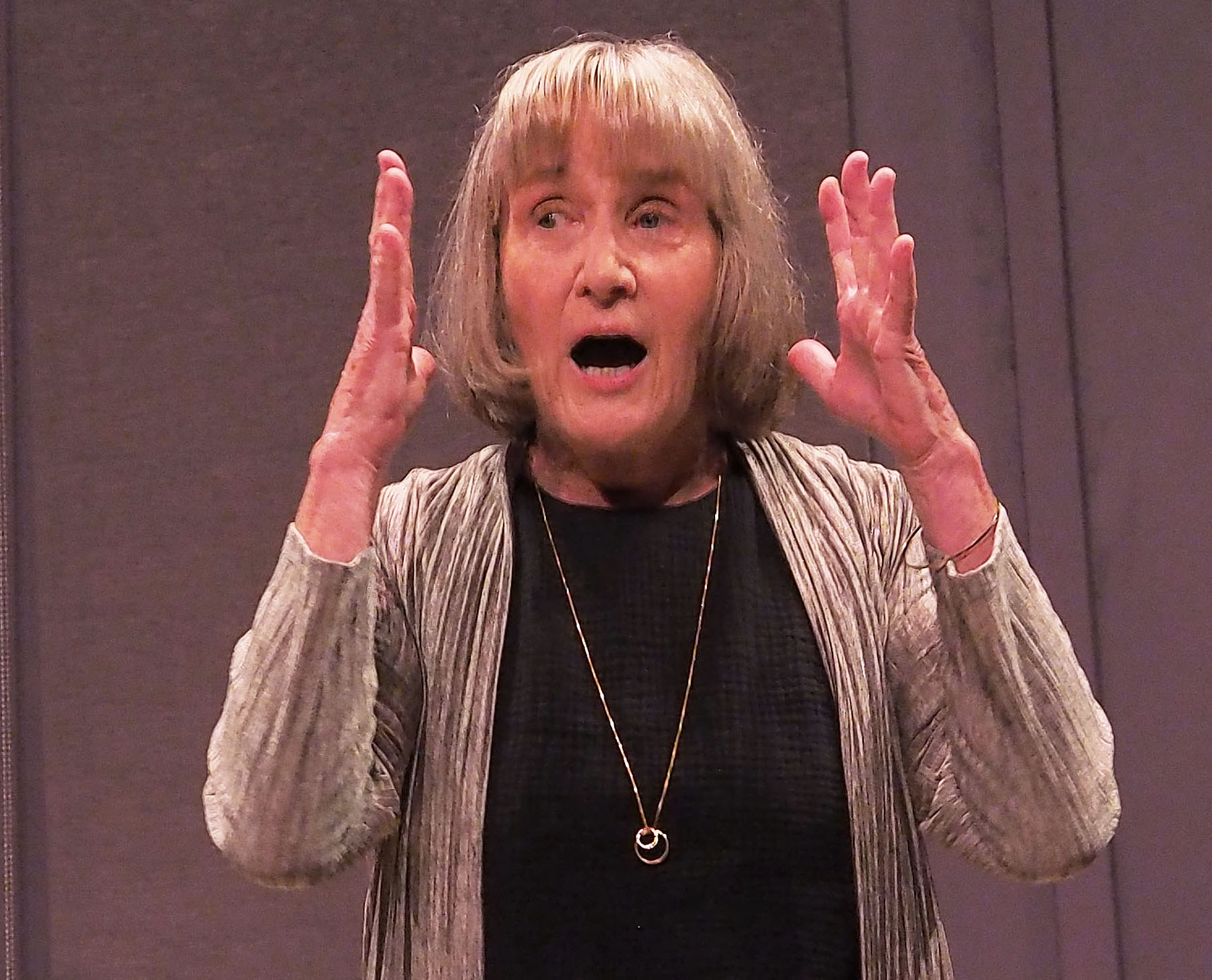 McBride, an older woman with a blonde bob haircut, holds her hands up with a surprised expression.