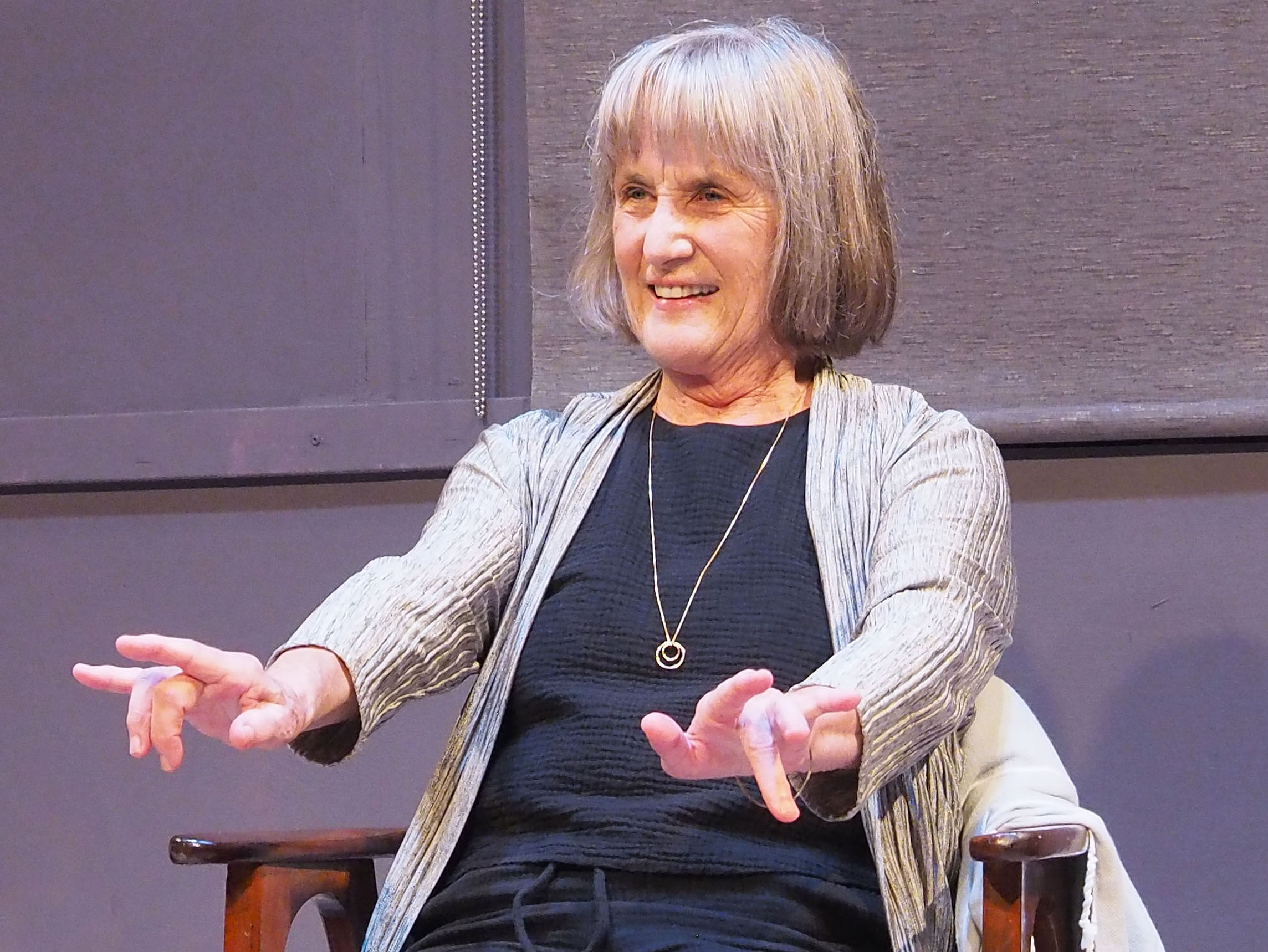 McBride, an older woman with a blonde bob haircut, looks out at the audience with a playful expression, miming typing or playing piano with her fingers.