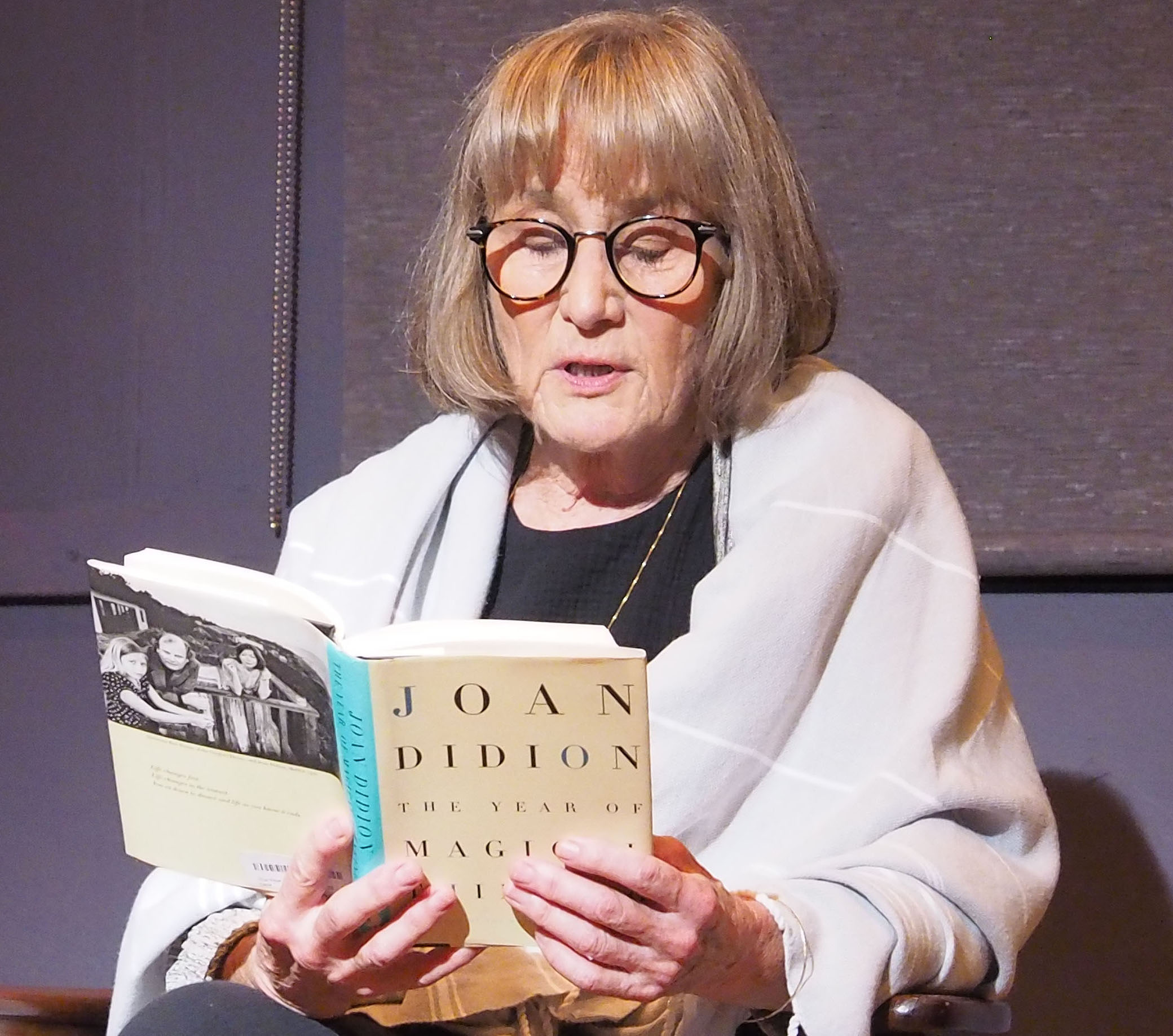 McBride, an older woman with a blonde bob, sits in a shawl reading American author Joan Didion's "The Year of Magical Thinking."