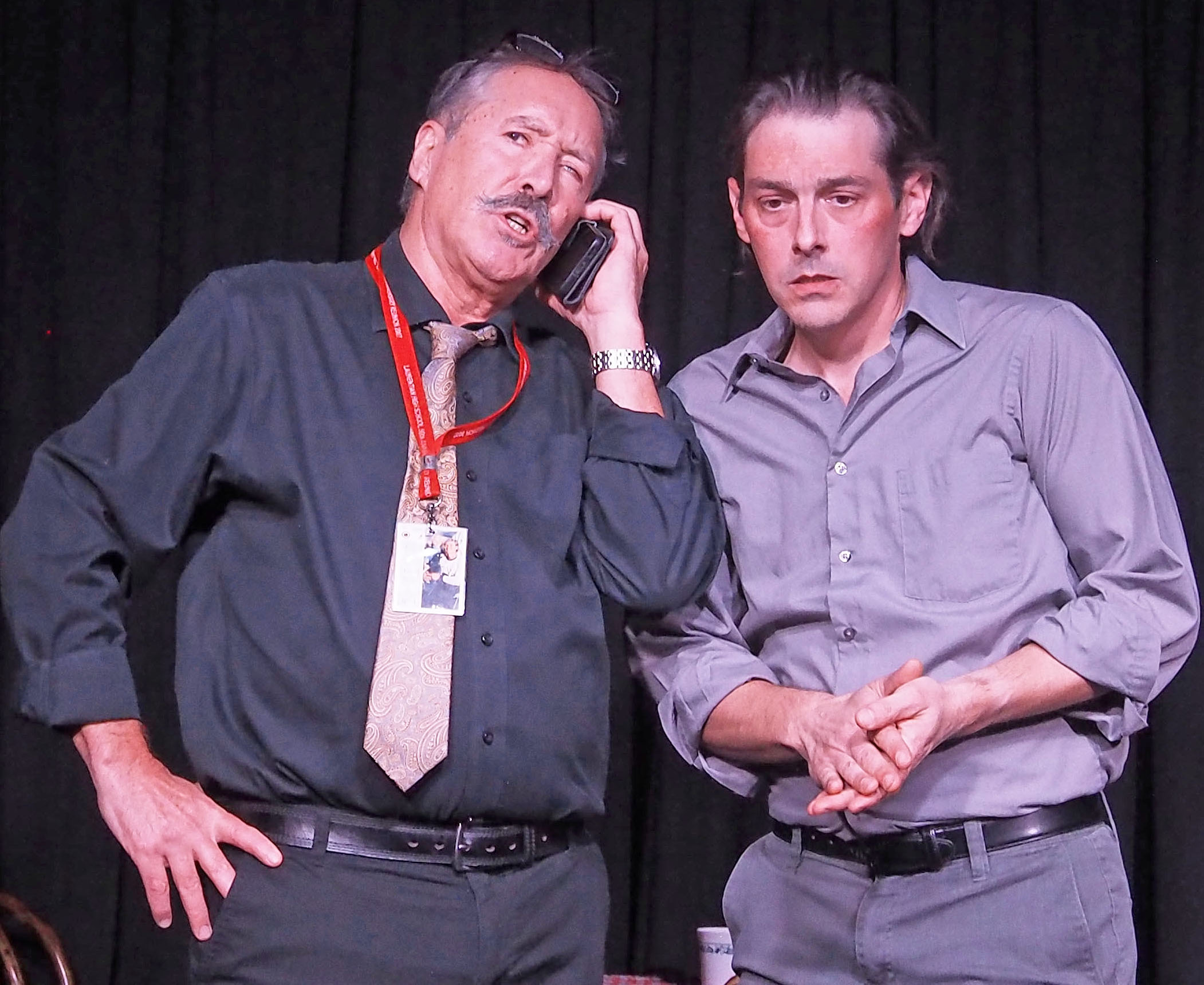 Two men stand facing the audience; the one on the left wears a red lanyard and talks on the phone, while the one on the right looks worried, with his hands clasped.
