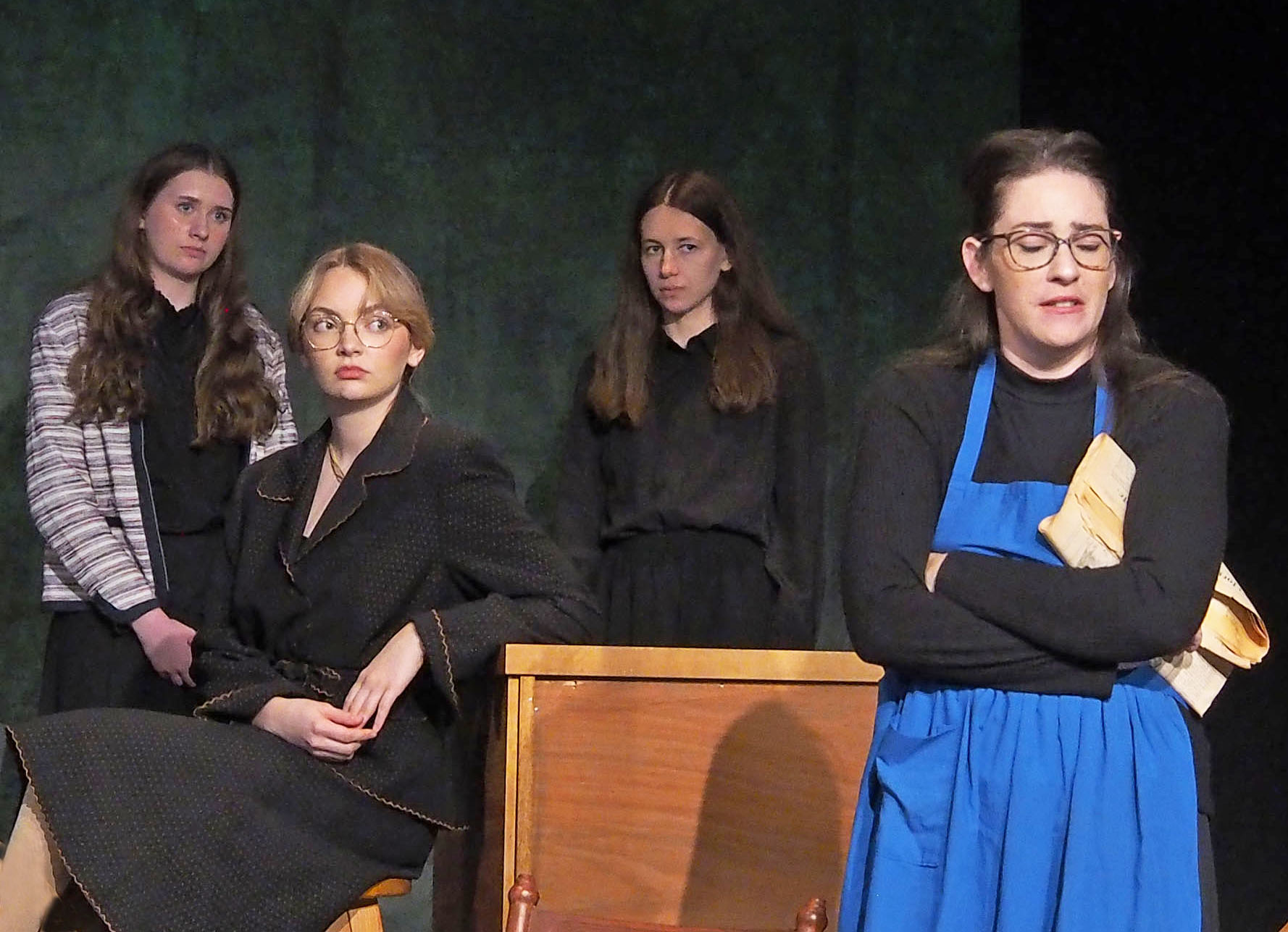 A group of actors stands before a dark backdrop wearing a range of suspicious and concerned expressions.