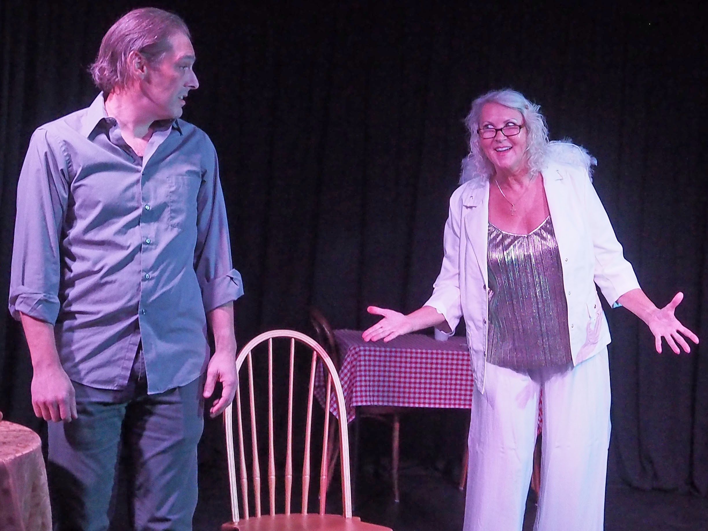 A man on the left looks over at a woman on the right in a white blazer, gesturing at him. Both face the audience.