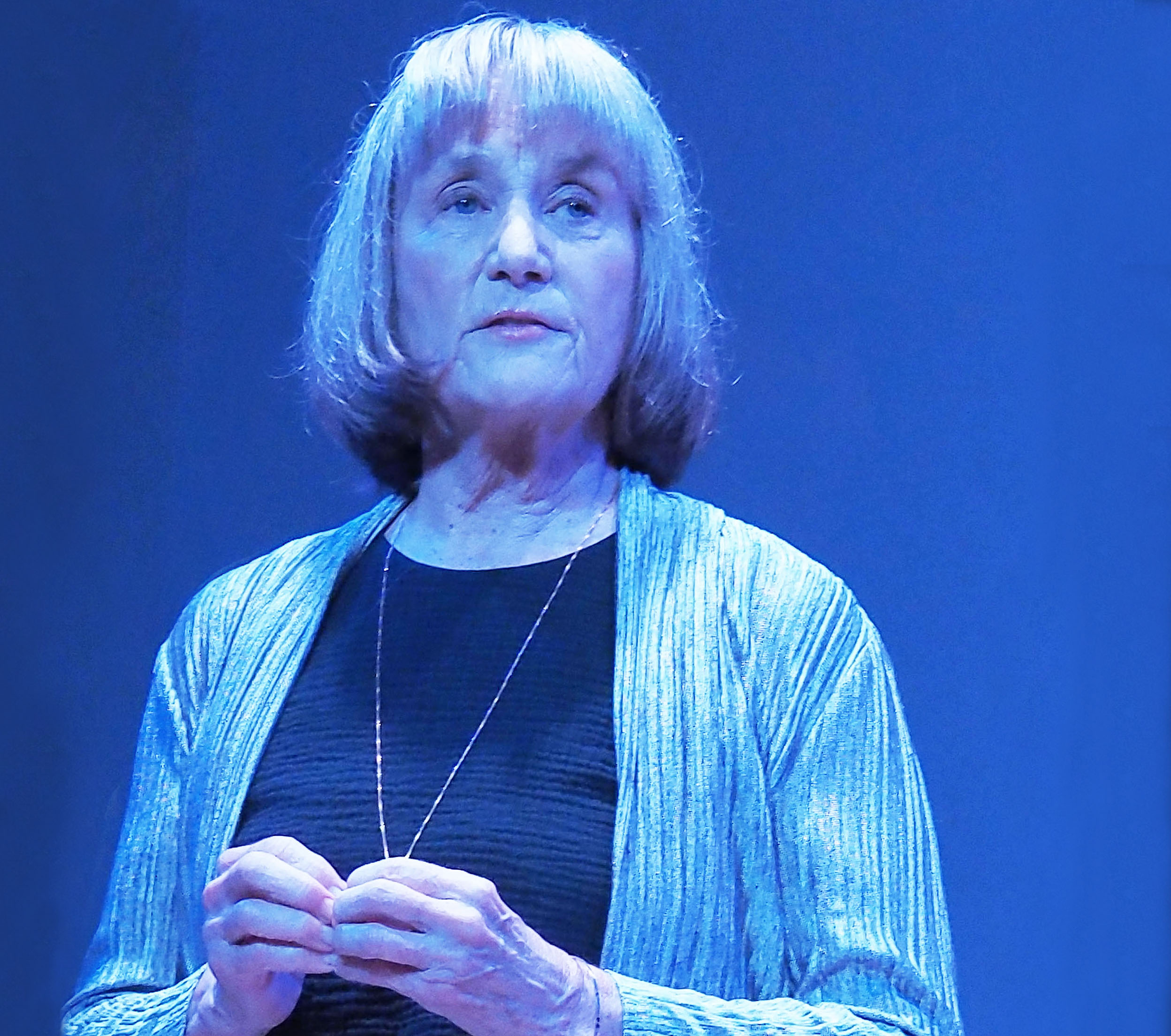 McBride, an older woman with a blonde bob haircut, stands in a blue spotlight looking into the distance.