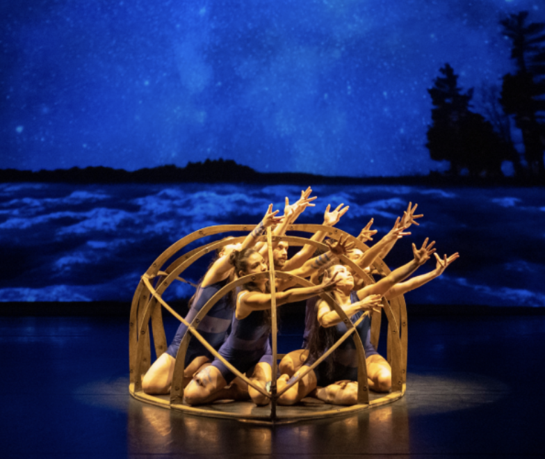 A group of dancers reach out of a hollow wooden structure and up to the sky, lit by a golden stage light and set against a background of the night sky.